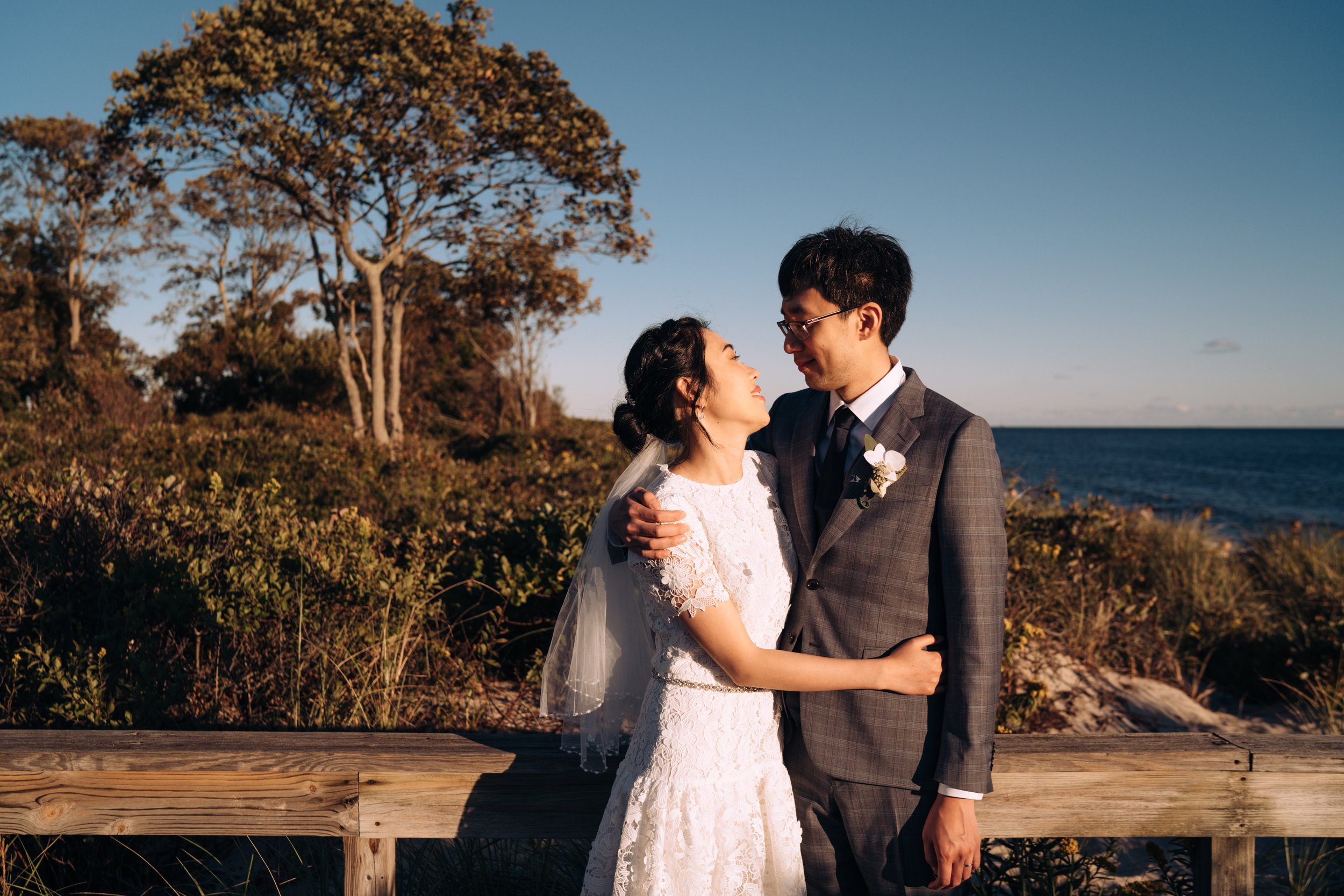 A Chinese couple, just married, stands side by side hugging and looking at each other against a blue sky and trees. The woman is wearing a short white dress and veil. The man is wearing a dark gray suit.