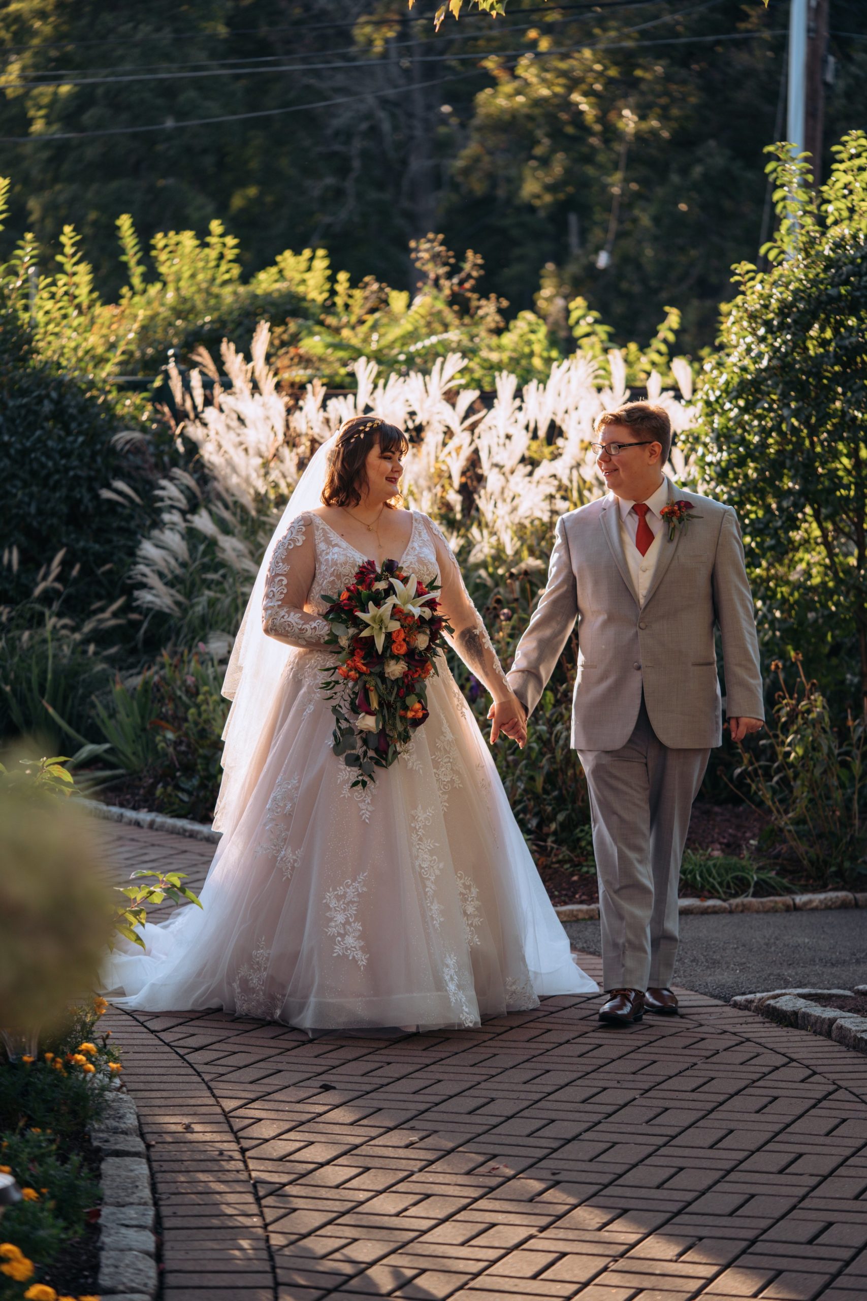 A white, cis-gendered woman in a wedding dress and veil holds hands with a white trans man in a gray suit. They are backlit and are walking toward the camera.