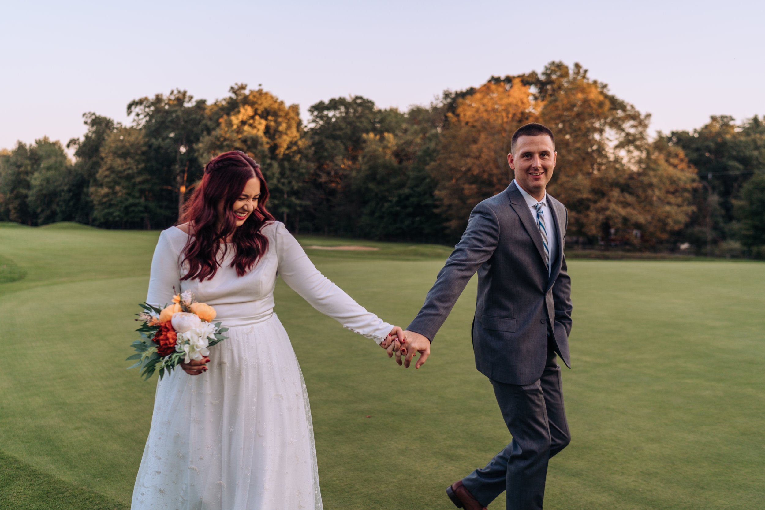 A new wife and husband hold hands and walk across a golf field, laughing.