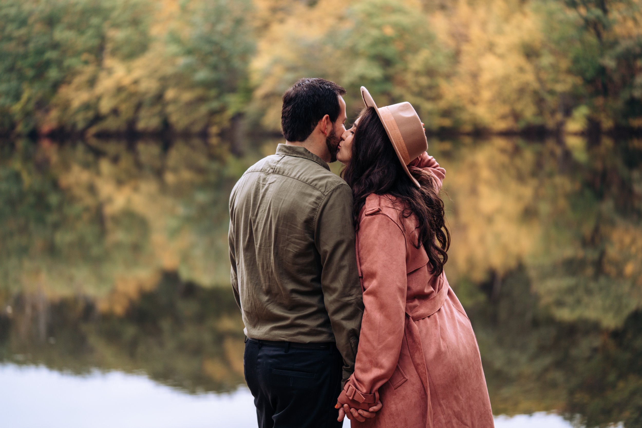 A couple stands against the backdrop of autumn trees reflected in a lake. They are both dark haired and are holding hands and kissing. The woman is wearing a pink trench coat and is holding a straw hat to her head with her free hand.