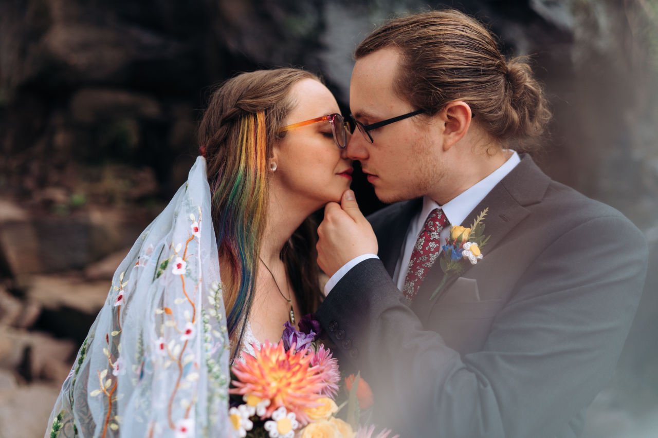 A bride with rainbow hair, a floral veil, and rainbow-colored glasses leans in for a kiss. Her groom has his hand under her chin and is wearing glasses, a suit, and a boutonniere.