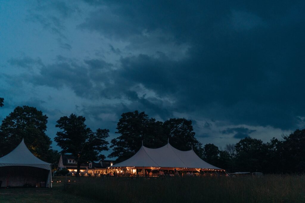 A nighttime photograph of a tented outdoor wedding at Smith Farm Gardens in Connecticut. The sky is dark blue with darker clouds; the trees in the background are nearly black; and the tent in the foreground is white with three spires and is lit with golden lights. It is summertime.