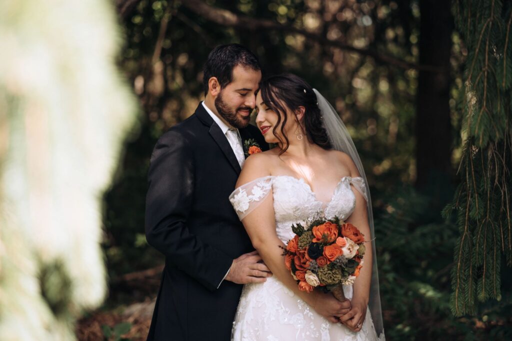 A bride and groom stand in the dappled light of a weeping evergreen tree. His hand is at her waist and she is holding a bouquet of dyed, wooden flowers. Their faces are touching.
