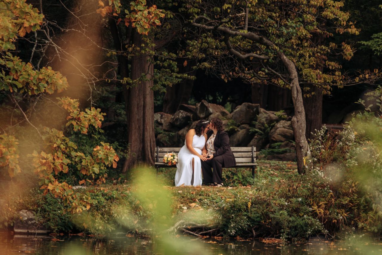 A lesbian couple sits on a bench holding hands as one partner kisses the other's forehead. One is wearing a white wedding dress and the other is wearing a black suit with a floral shirt underneath. They are framed by trees and foliage.