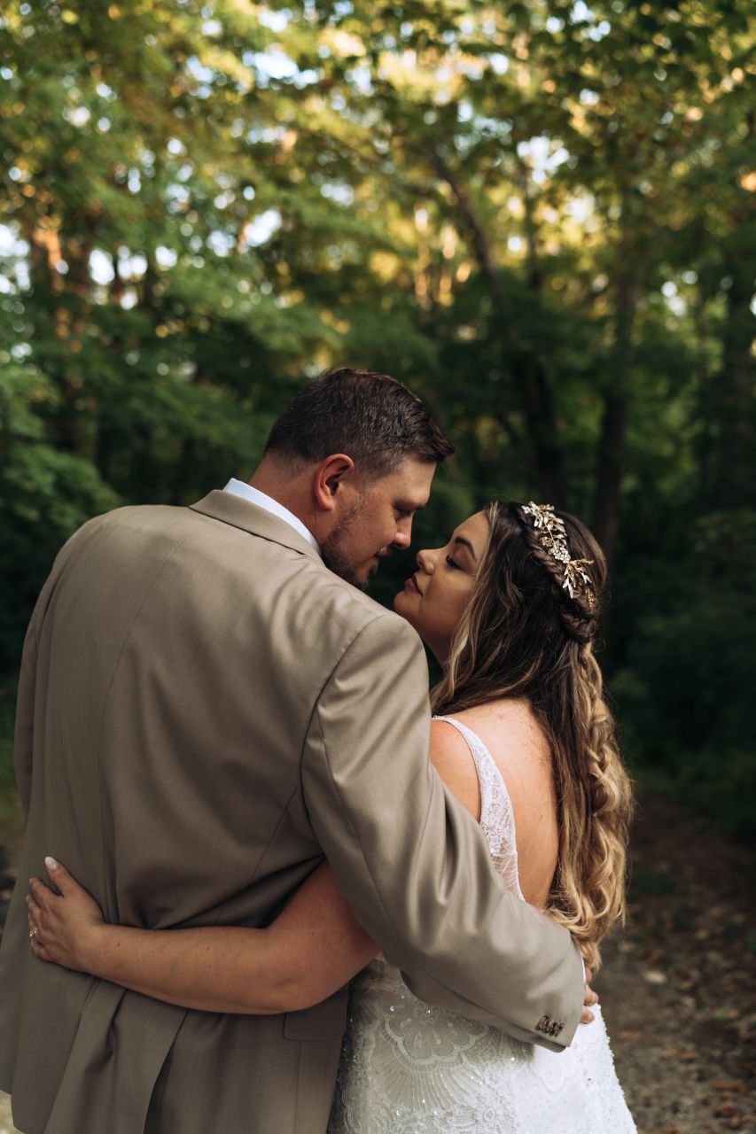 A groom, a tall white man wearing a tan suit, has his arm around a bride, a white woman with long hair who is wearing a white wedding dress and a tiara. They are facing away from the camera and are almost touching noses.