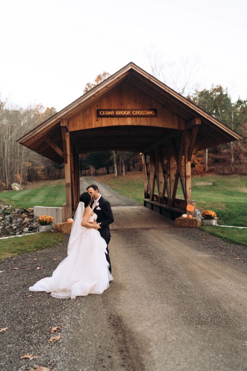 Bride in a dreamy, full wedding gown and groom in a classic suit kiss near a covered bridge in Connecticut.