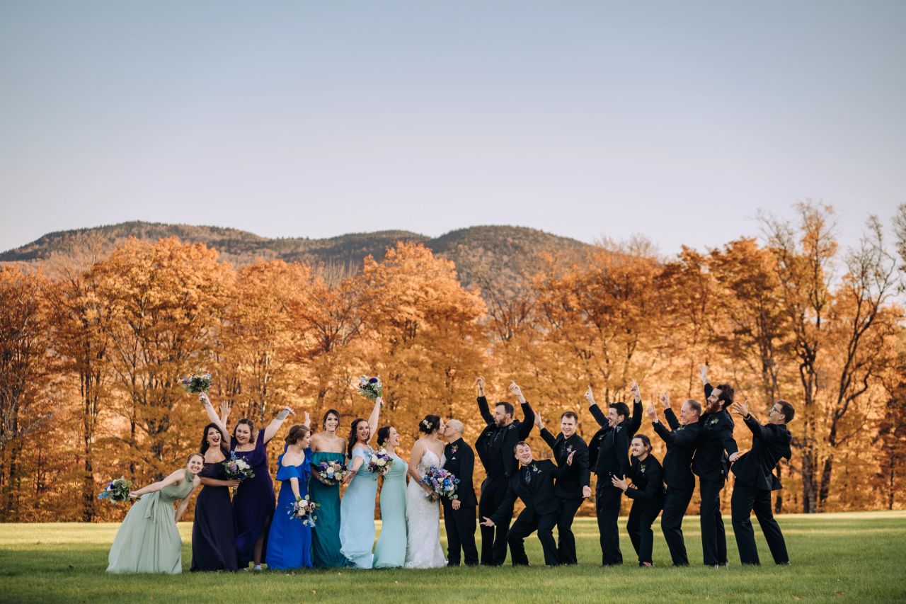 Wedding party in jewel tones cheers in front of Vermont mountain in the Fall.