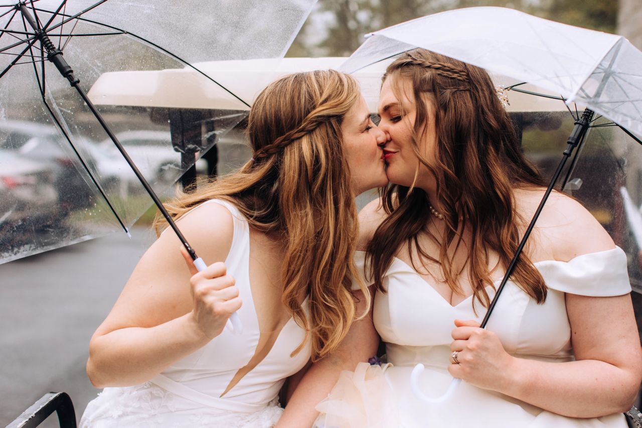 Two brides, both in white dresses, sit and kiss. They are each holding an umbrella, as it is raining.