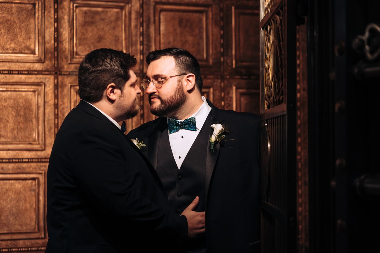 A newlywed couple, two grooms, both in tuxes, face each other with quiet faces. One man has his hand on the other's vest.