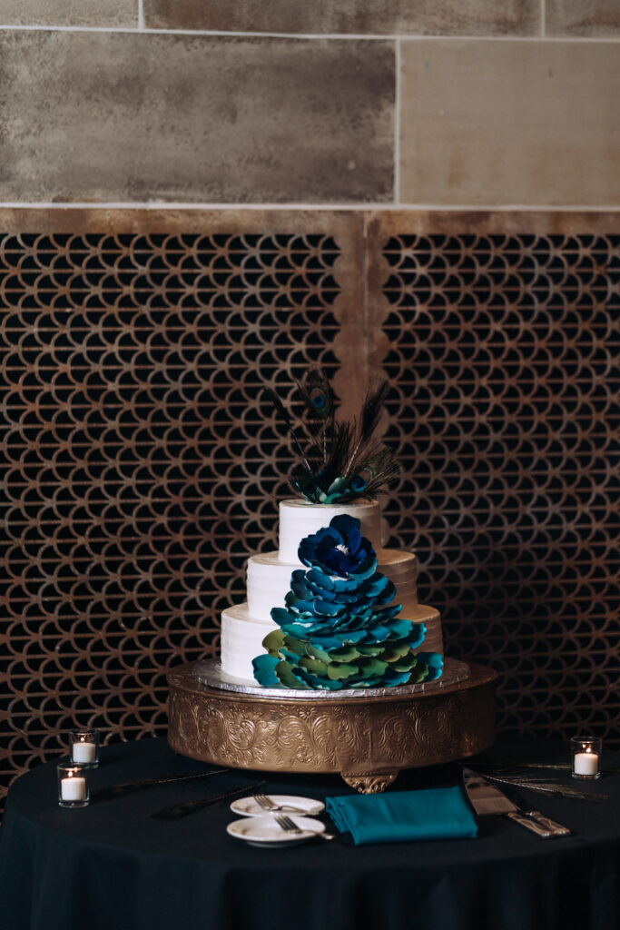 A three-tiered white wedding cake decorated with jewel-toned blue and green flower petals and a peacock feather.