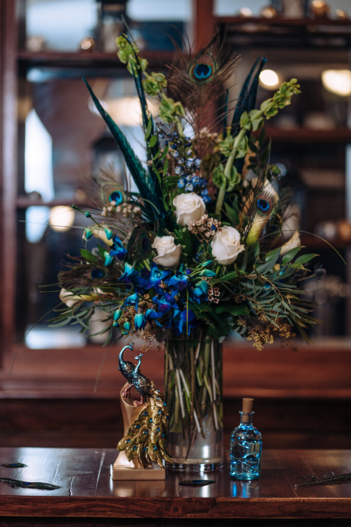 A tall, dramatic wedding floral arrangement with peacock-themed colors, peacock feathers, and white roses.