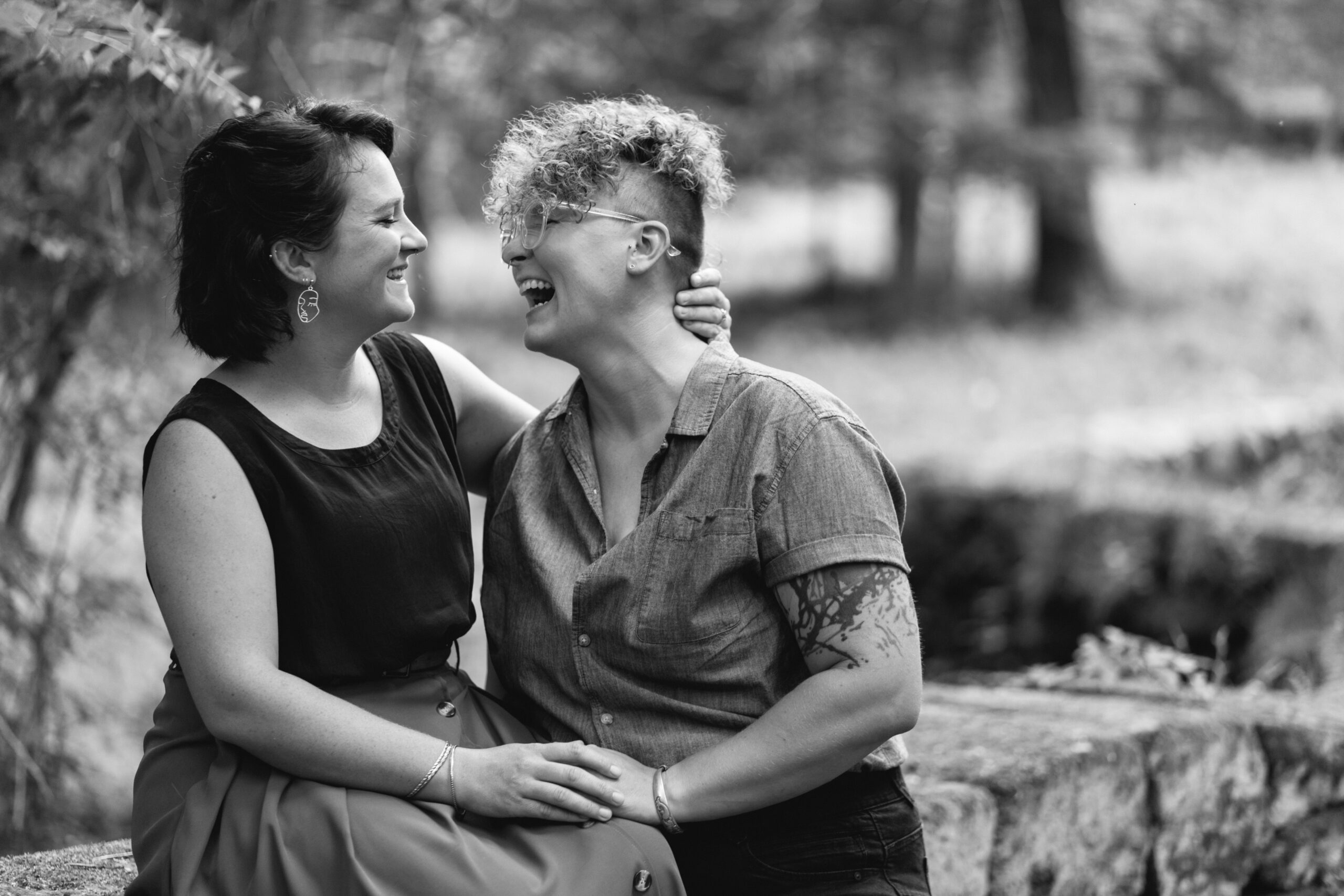 A lesbian couple, one sitting on a stone wall and the other standing, holding her hand, looks at each other and laughs. The image is black and white.