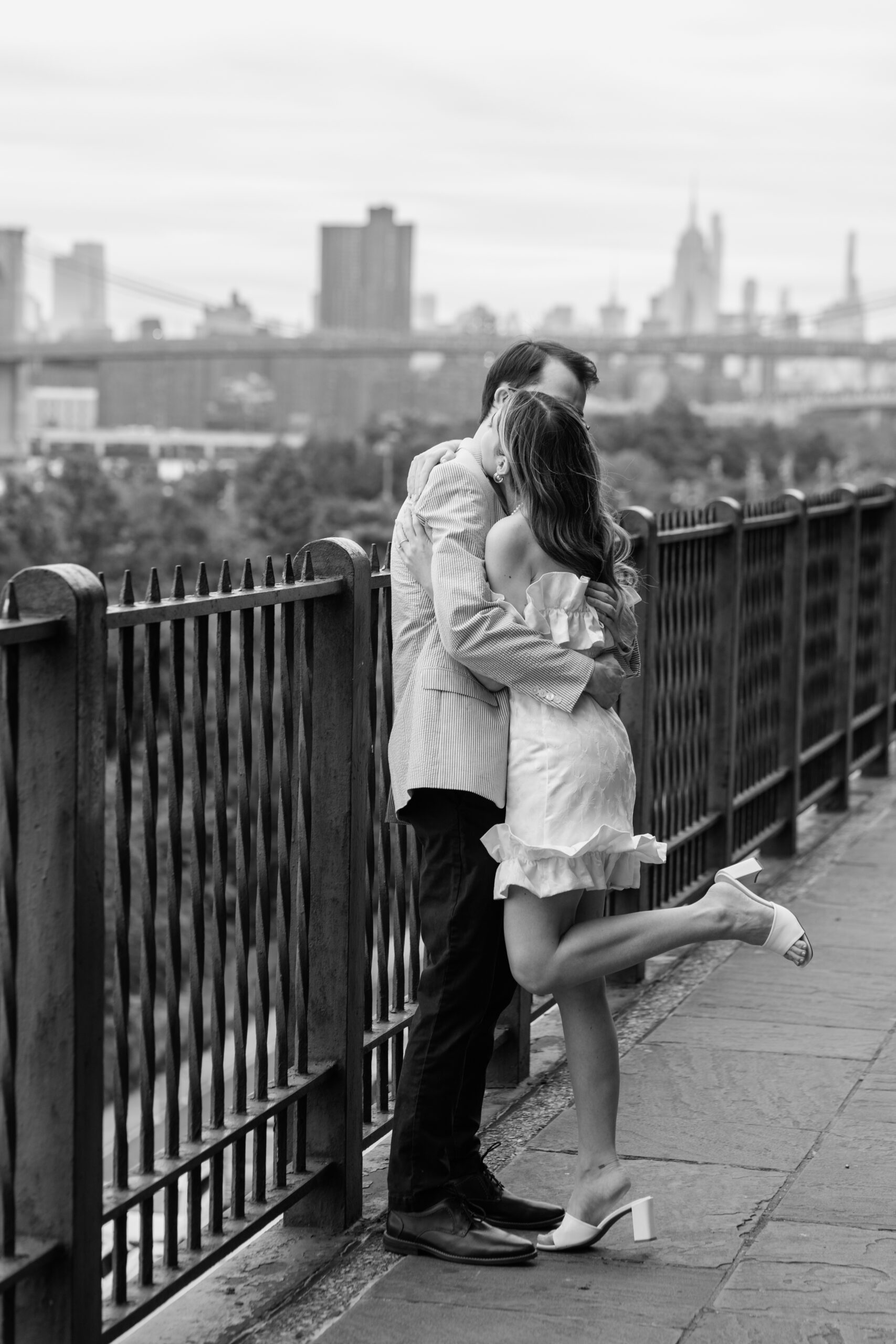 An engaged couple--a thin white man in dark pants and a seersucker suit jacket and a thin white woman in a stylish short white dress and heels hug on the Brooklyn Heights Promenade. She is kicking up her heel. The photo is black and white.