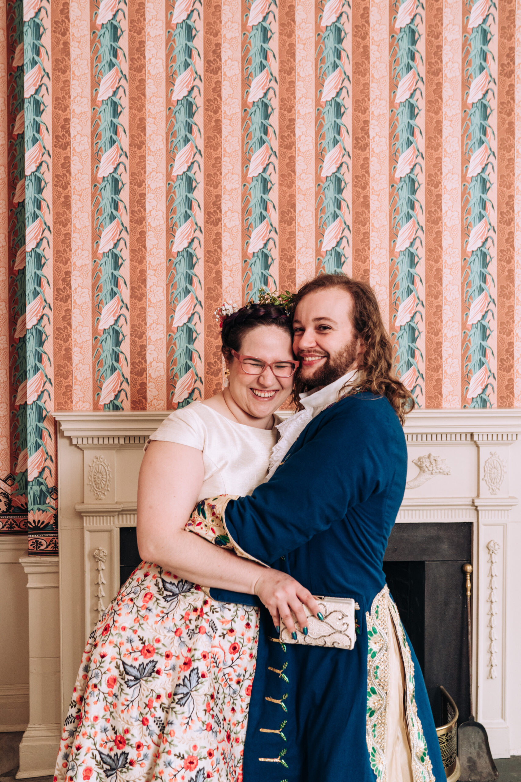 A queer couple Maria (she/her) and Naomh (he/him) hug and laugh on their wedding day. They both wear bespoke Renaissance outfits and are standing in front of bold, striped wallpaper.